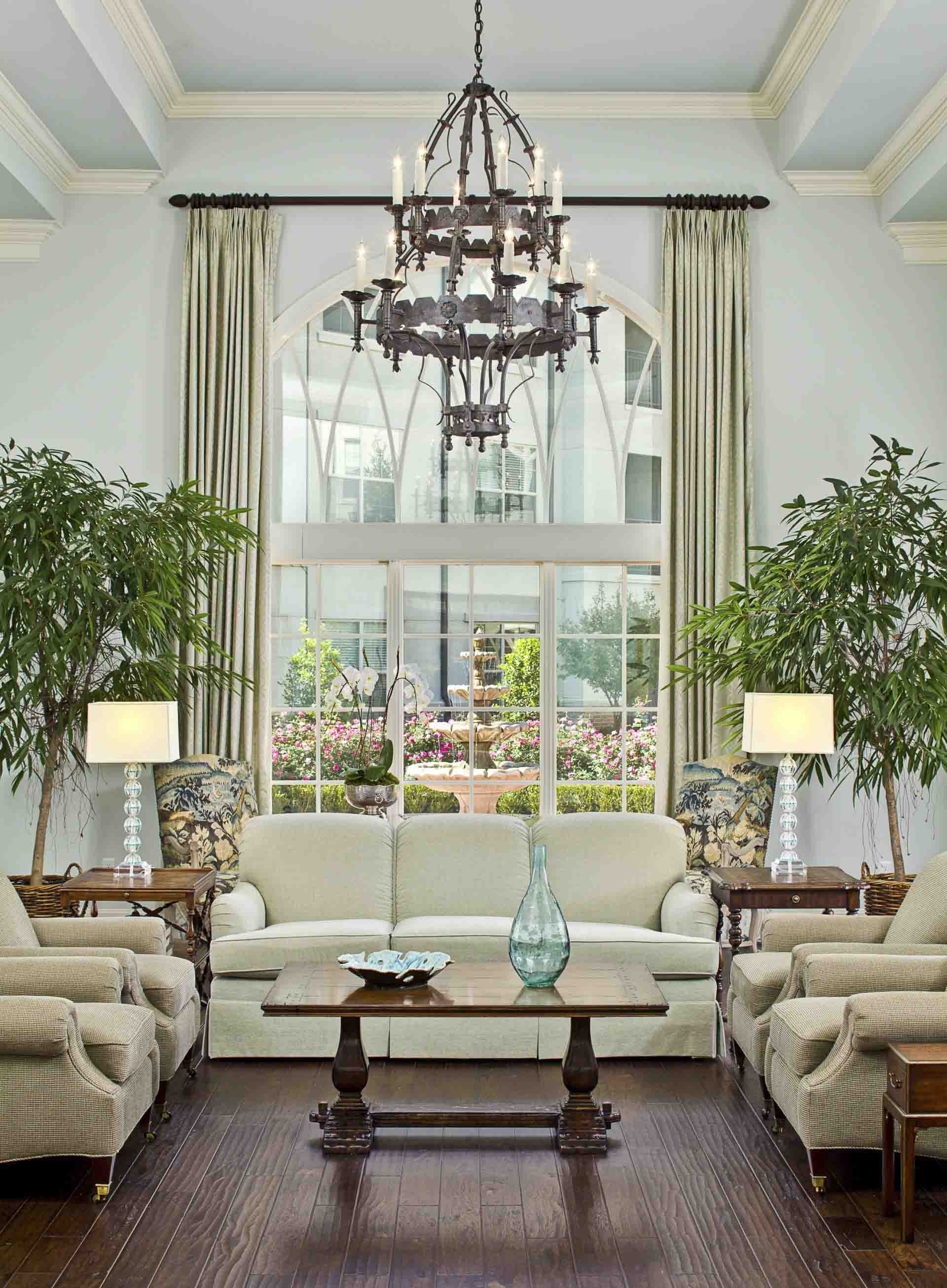 A sitting area with chandelier & large window overlooking a courtyard at Tradition Senior Living - Prestonwood in Dallas, TX.