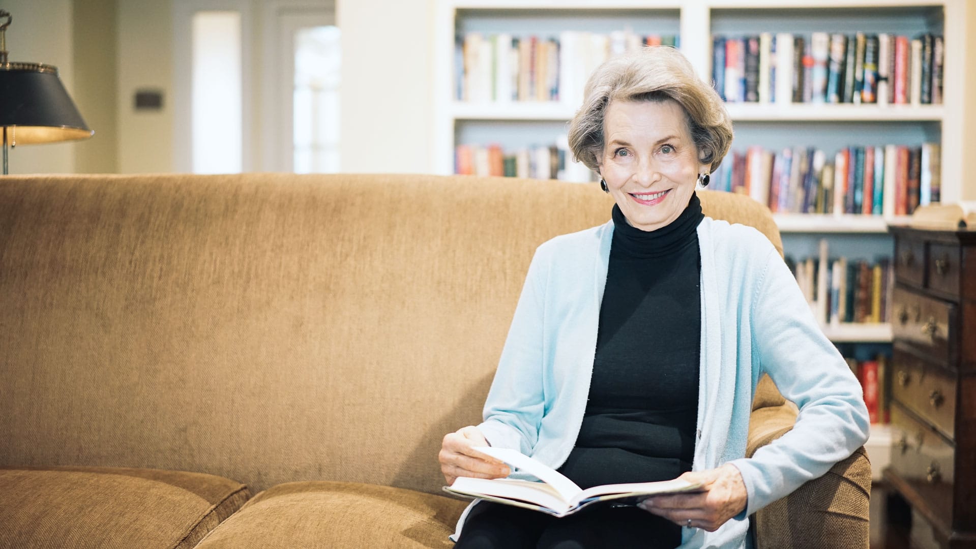 A woman sits on a couch & reads a book at the library inside Tradition Senior Living in Dallas, TX.